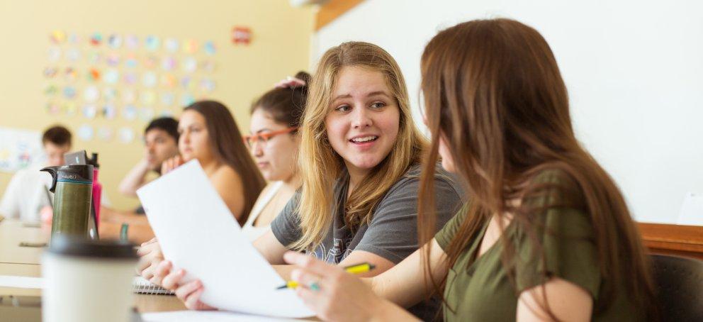 Two students in class are talking to each other while holding paper and pens